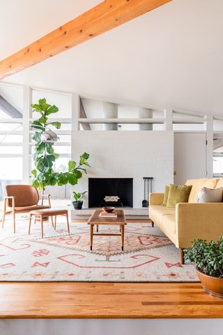 A living room by Urban Allure with central fireplace, tan leather and wooden chair with matching footstool, yellow sofa, large rug