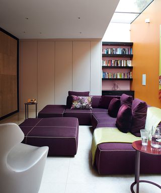 A large purple modular sofa and matching footstool in a modern white and orange living room with skylight.