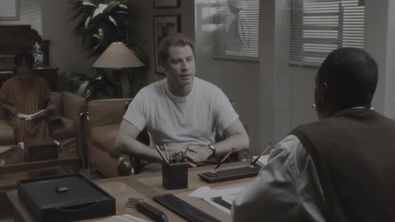 John Travolta with blonde hair speaking to a man at a desk.