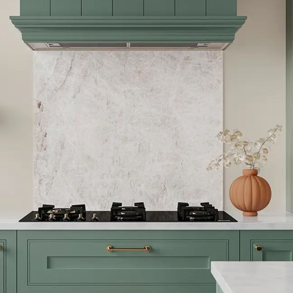 A sage green shaker kitchen with brass handles and a matching panelled chimney hood, featuring a large slab of soft cream and blush quartzite as a splashback behind a black four-burner gas hob. A terracotta ribbed vase with dried stems sits on the white worktop alongside.