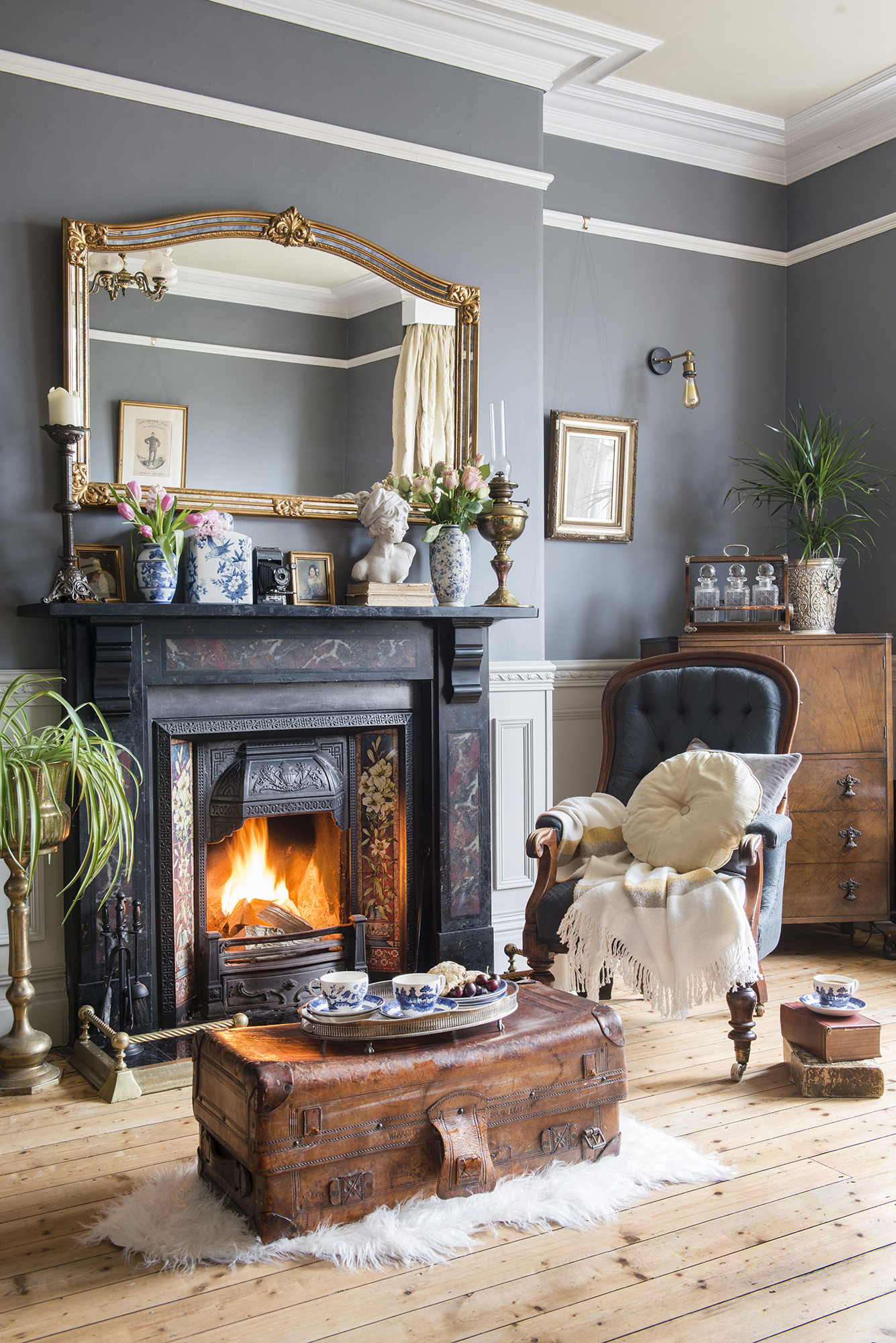 A grey living room with renovated Victorian home fireplace, large mirror over mantel and set of whiskey decanters in makeshift home bar within alcove area