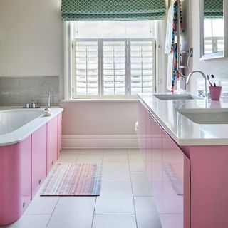 a pink bathub with matching pink undersink cabinets with double inbuild sinks, and a window with a green and brown pattern blind