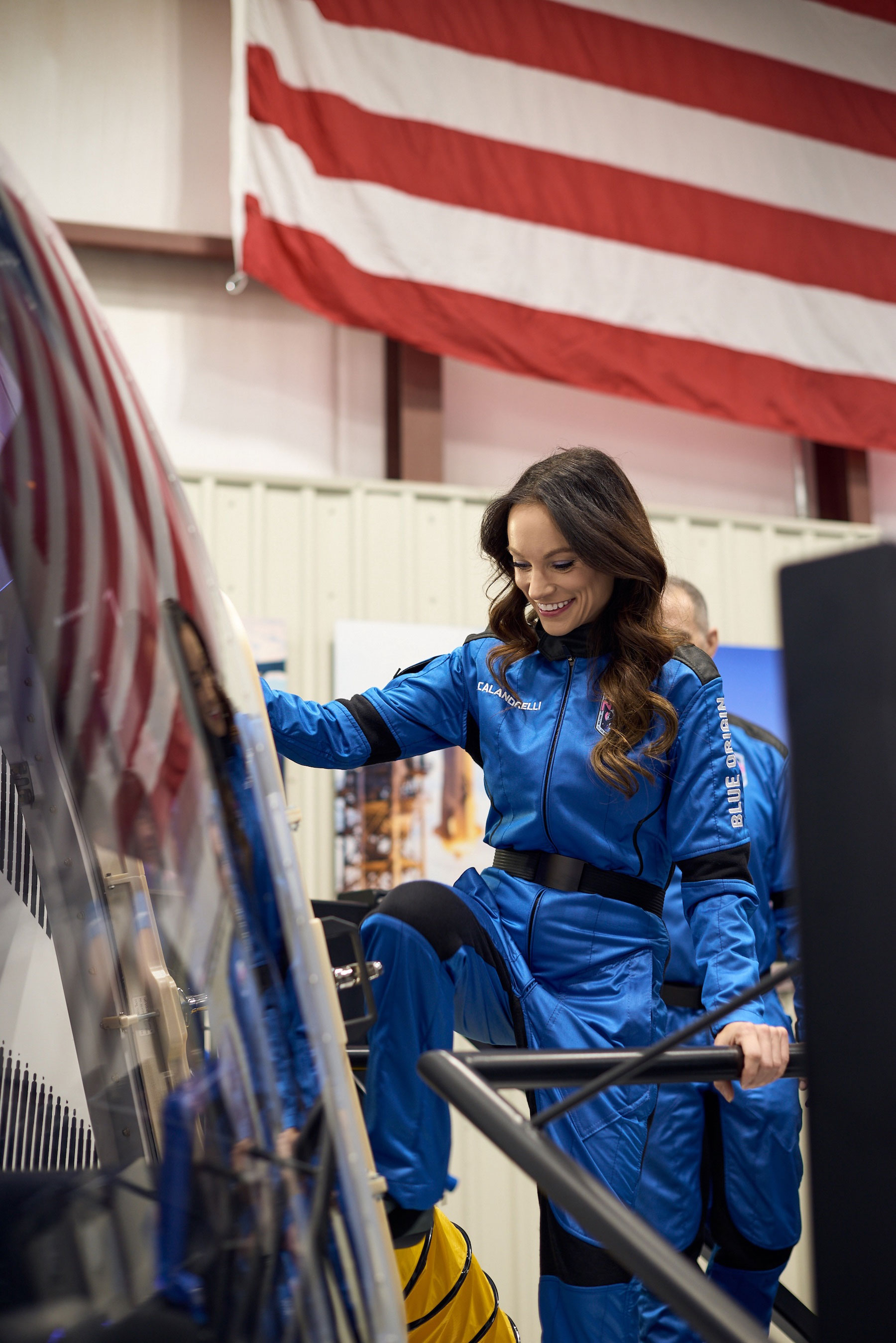 a woman in a blue flight suit enters a spacecraft inside a hangar