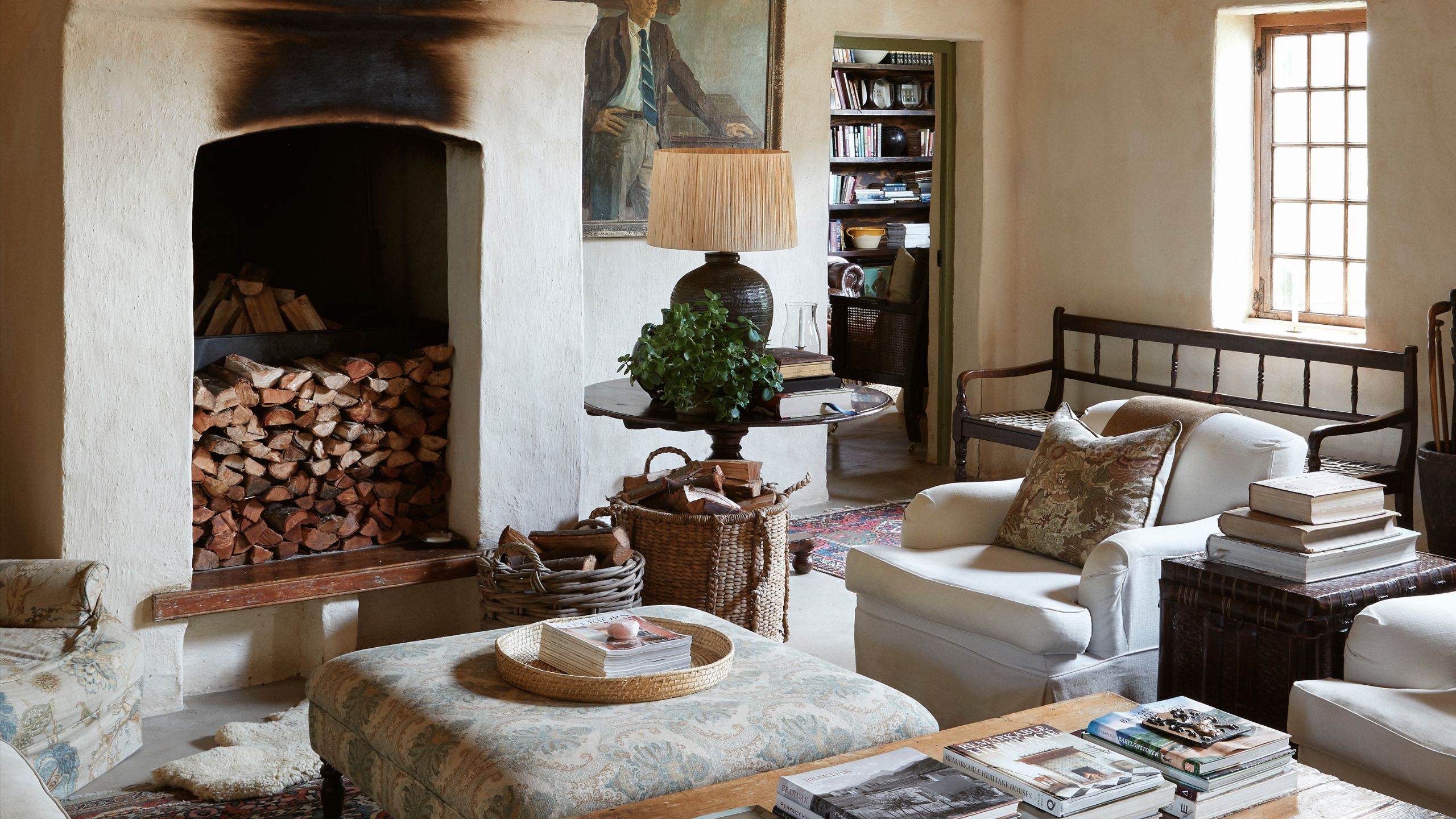 Cozy rustic living room featuring a large hearth filled with stacked firewood, cream armchairs, a patterned ottoman coffee table, and an adjacent library.
