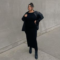 gia wears black dress, black boots, and black jacket while posing outside. 