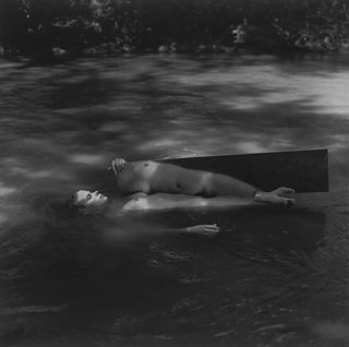 A nude woman lying on her back in a stream holding a long mirror.