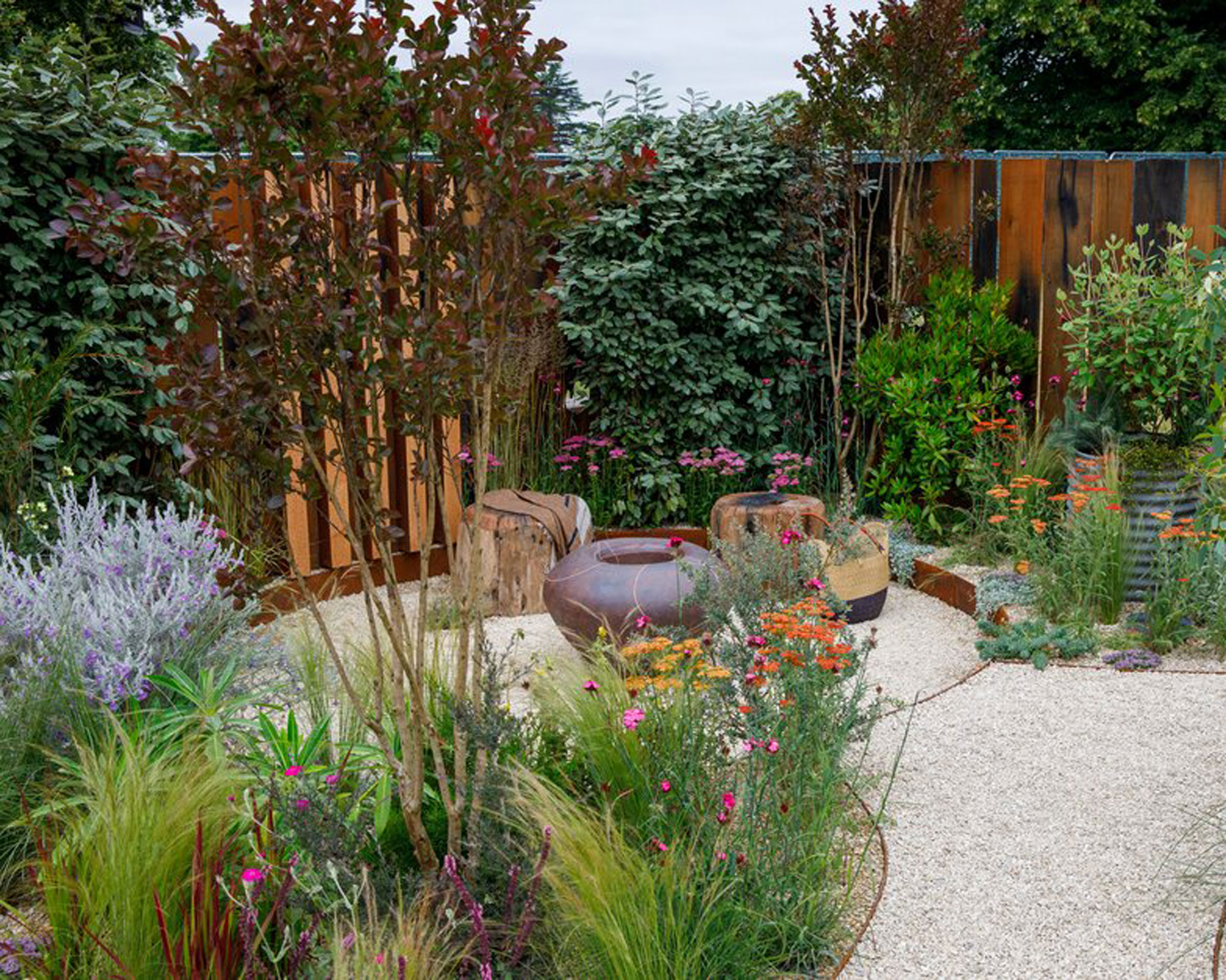 modern garden design with fence, firepit, garden seats, gravel landscaping and deep flowerbed with plants and ornamental tree