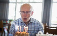 An man with white hair and glasses blows out candles on a birthday cake.