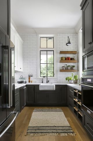 monochromatic kitchen with wooden shelving on the right of window with spice jars and plants, white metro wall tiles, black cabinetry, hardwood floor, rug