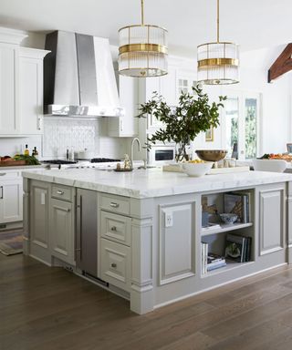 Large kitchen space with large square kitchen island with gray cupboards, draws, shelves, white marble countertop, dark wood flooring, two glass and gold large pendants hanging over island, white kitchen cabinets