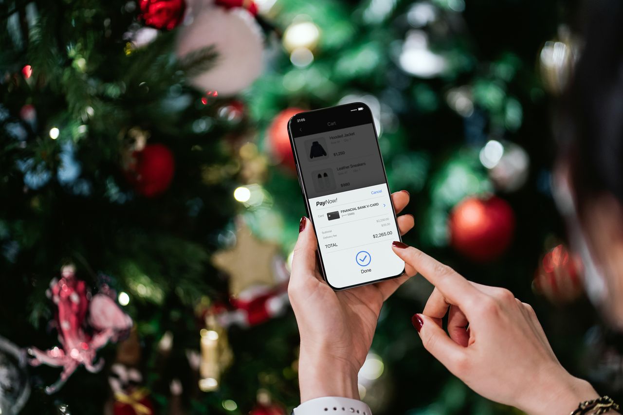 Over the shoulder view of woman&#039;s hand purchasing Christmas gift online with smartphone and making mobile payment with credit card against illuminated Christmas tree. 