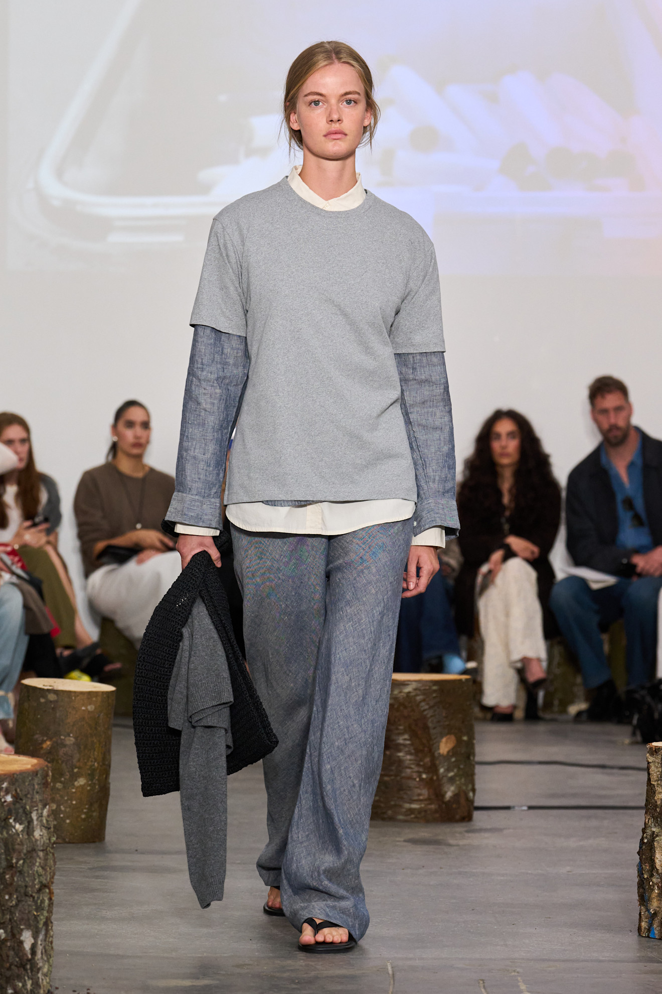Layering staples for spring 2026 is shown in a photo from Aiayu's runway show, with a model walking while wearing a white button-down layered underneath a blue button-down and gray t-shirt styled with blue wide-leg pants, black flip-flops, and extra sweaters 