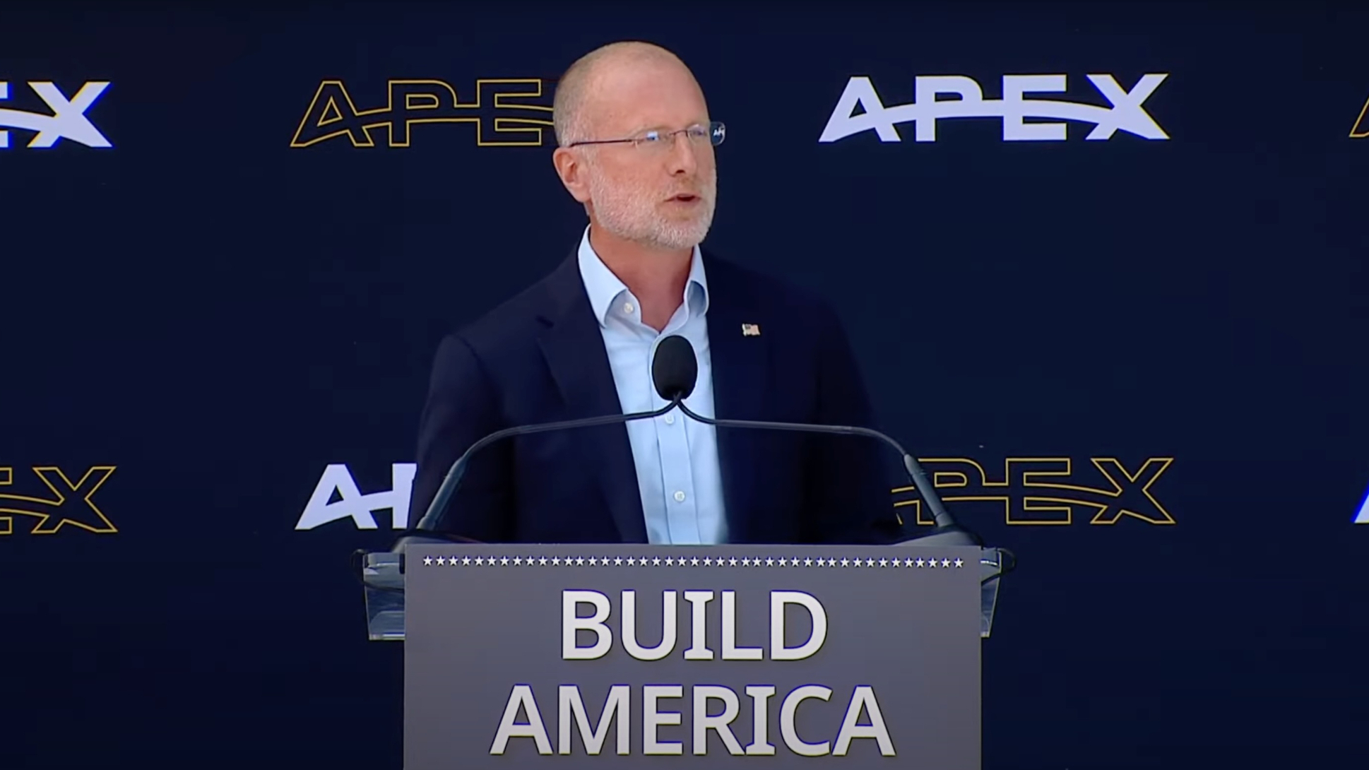 an older bald man with glasses wearing a blue blazer speaks at a podium with the words &quot;build america&quot; on it