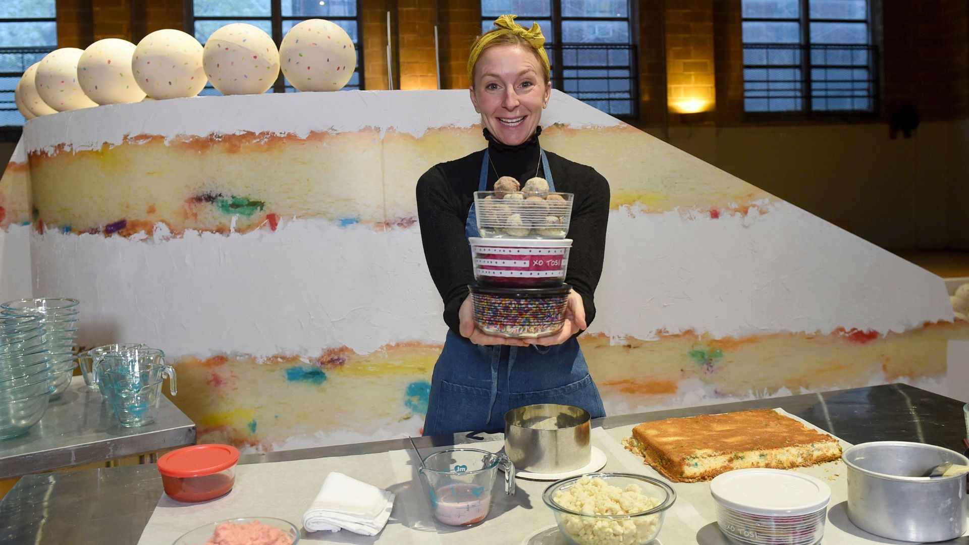 Christina Tosi holds some Tupperware in front of a giant funfetti cake