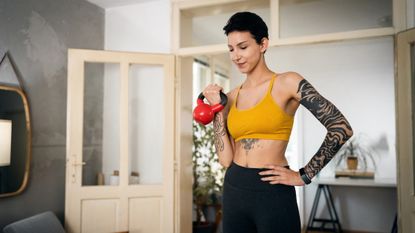 A tattooed woman in a yellow crop top does a bicep curl with a red kettlebell. She is in a home setting and looks down, away from the camera.