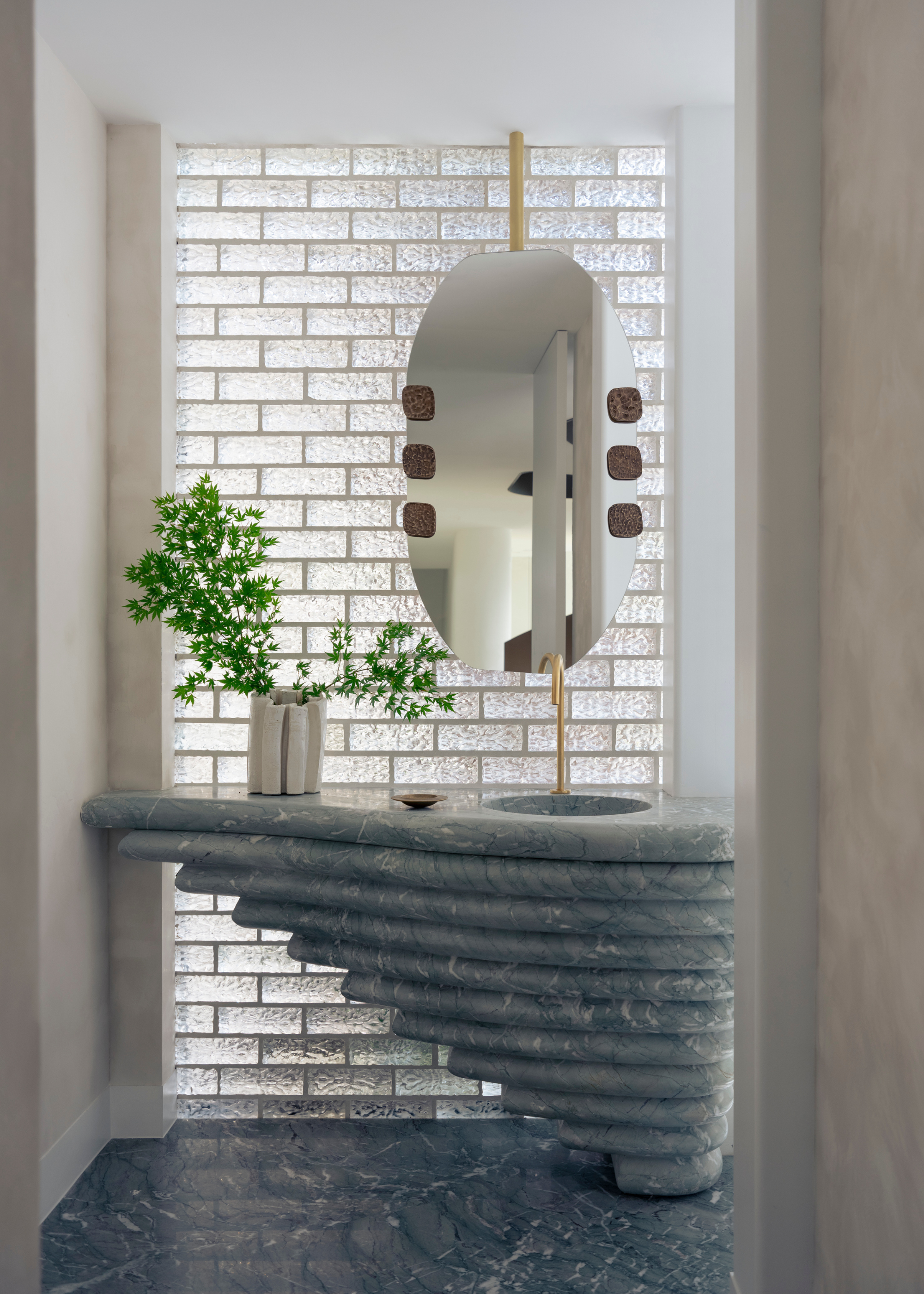 A bathroom with a stacked marble sink, a vase of green stems, a hanging mirror, and a textured glass brick wall
