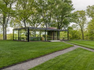 Day time image of Phillip Johnson’s Glass House, lawn with gravel pathway leading to the building, surrounding trees and landscape