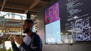 A man giving a presentation in front of a screen showing overlapping windows of python code