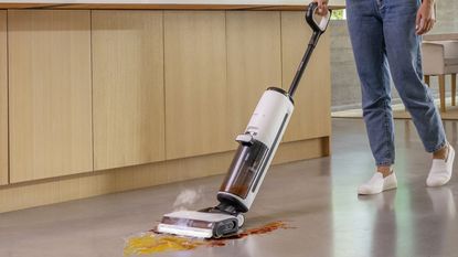 A woman wearing dark denim jeans and white footwear cleaning a kitchen spill using the Tineco Floor One S7 steam wet-dry vacuum 