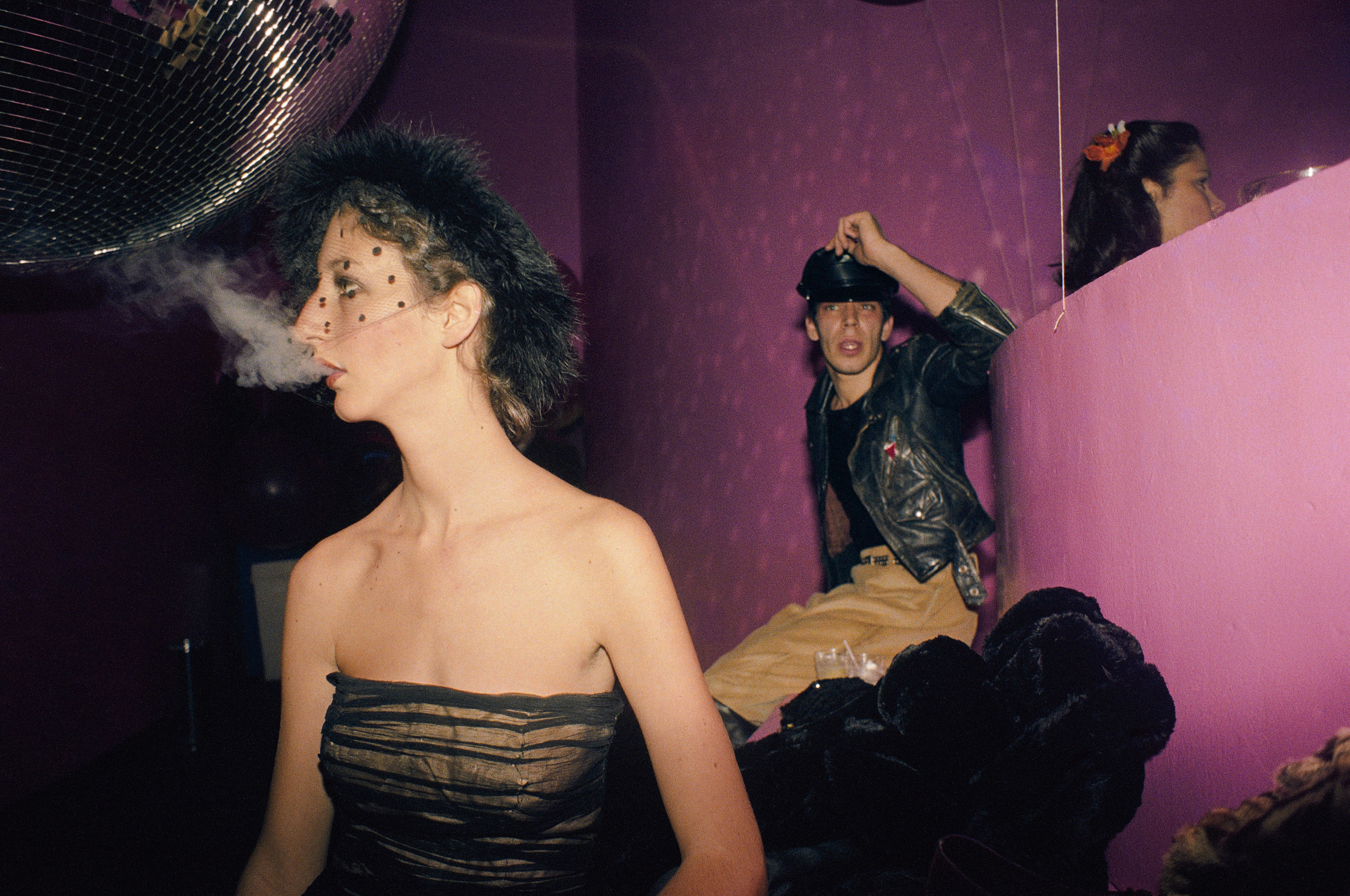 In a dimly lit room with purple walls and a disco ball, a woman in a sheer black top exhales a cloud of smoke while a man in a leather jacket sits behind her.