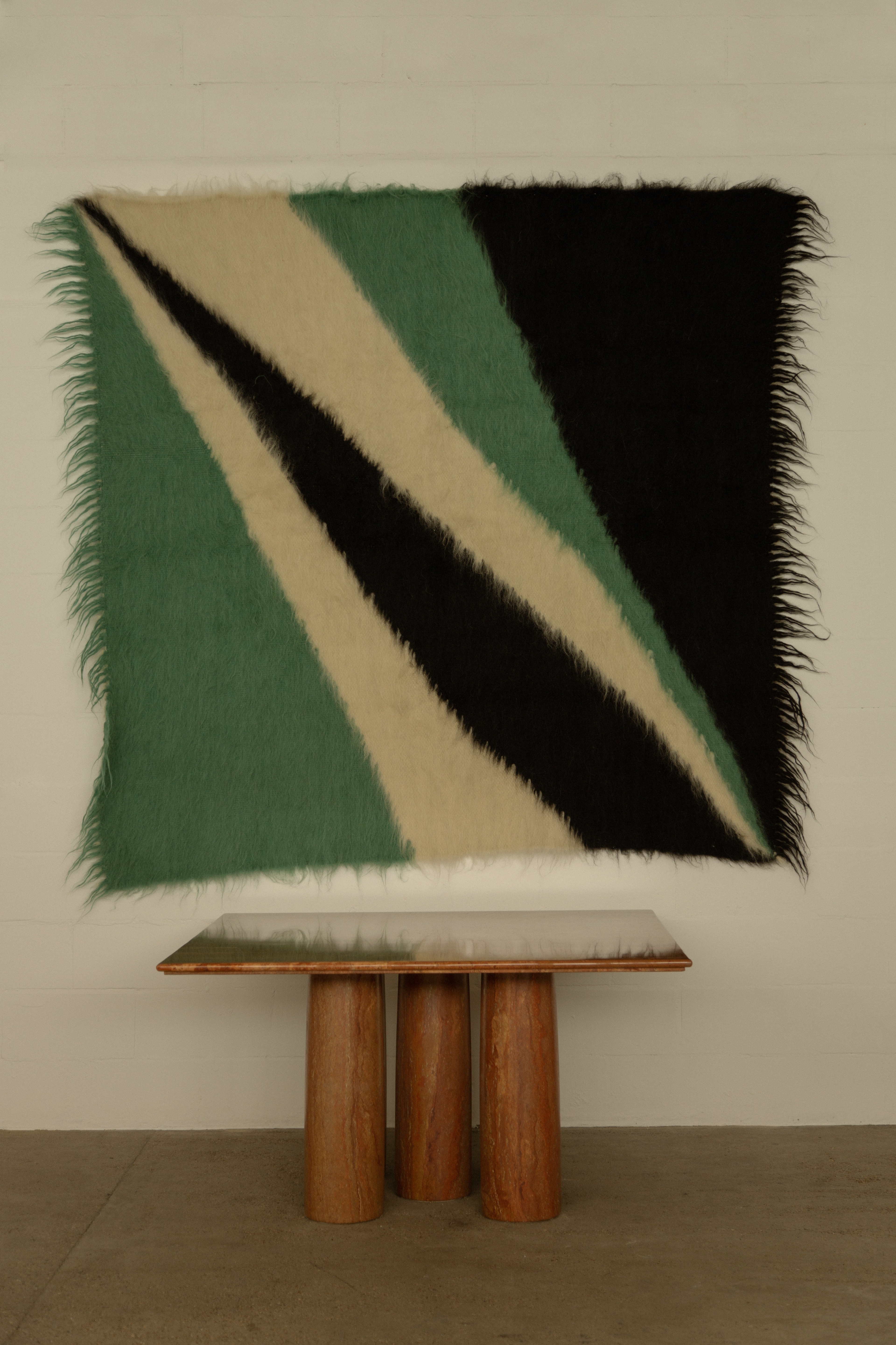 A colorful, abstract textile tapestries in swathes of green, beige, and black hangs atop a wooden coffee table with three chunky legs.