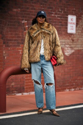 A woman standing on a sidewalk at New Fashion Week wearing distressed wide-leg jeans, a white tee shirt, fur coat, heels, and hat