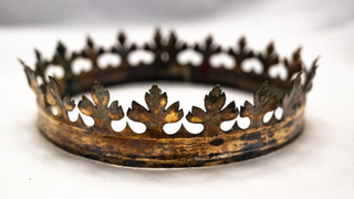 A tarnished gold crown ringed with fleur-de-lis type flowers sits on an off-white piece of fabric