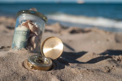 glass jar with money and old gold compass lying in the sand on the beach for retirement destination