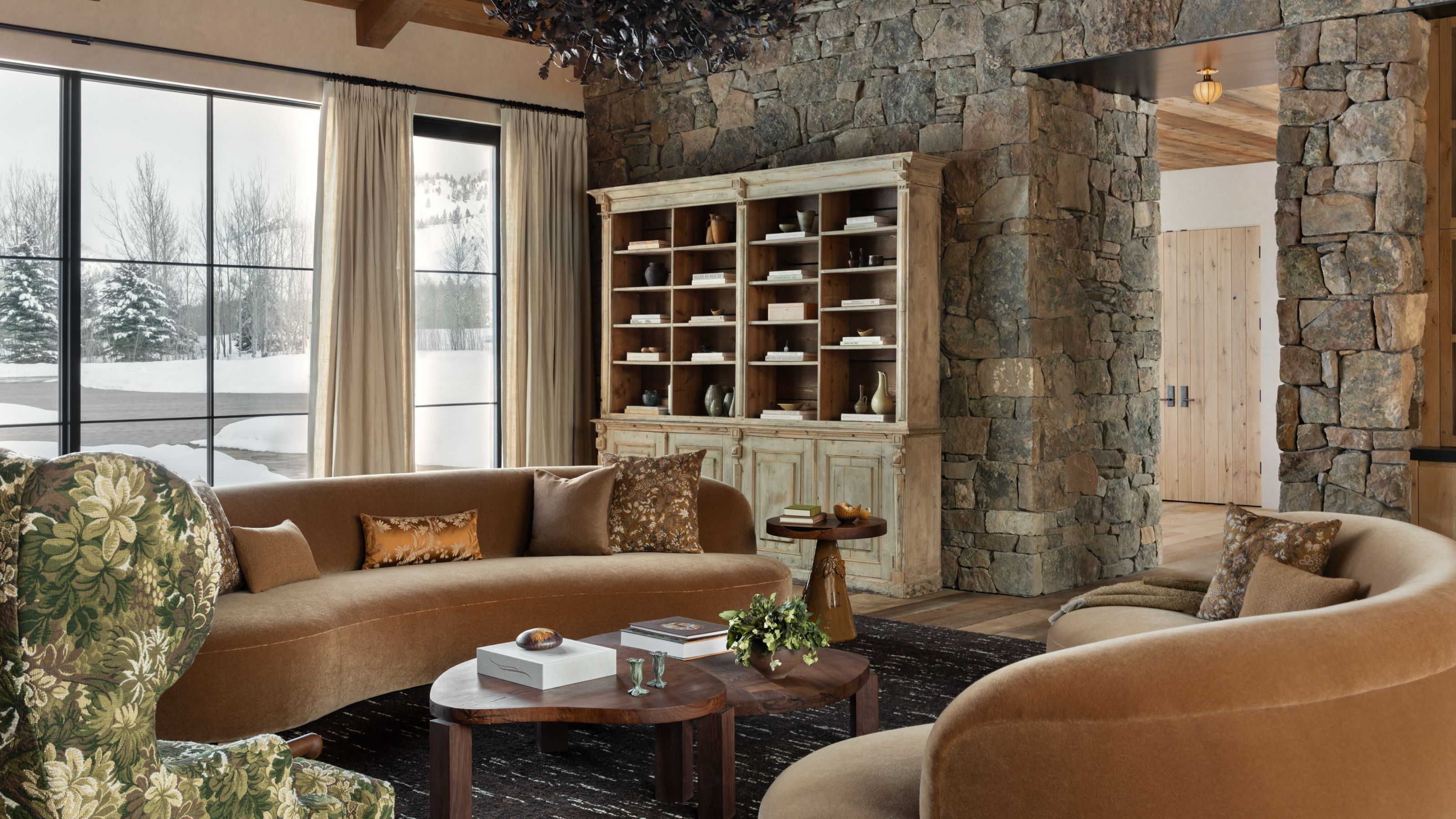 a stone clad living room with an open arched door, large glass doors looking out to a snowy yard, a rustic bookcase, with curved velvet couches