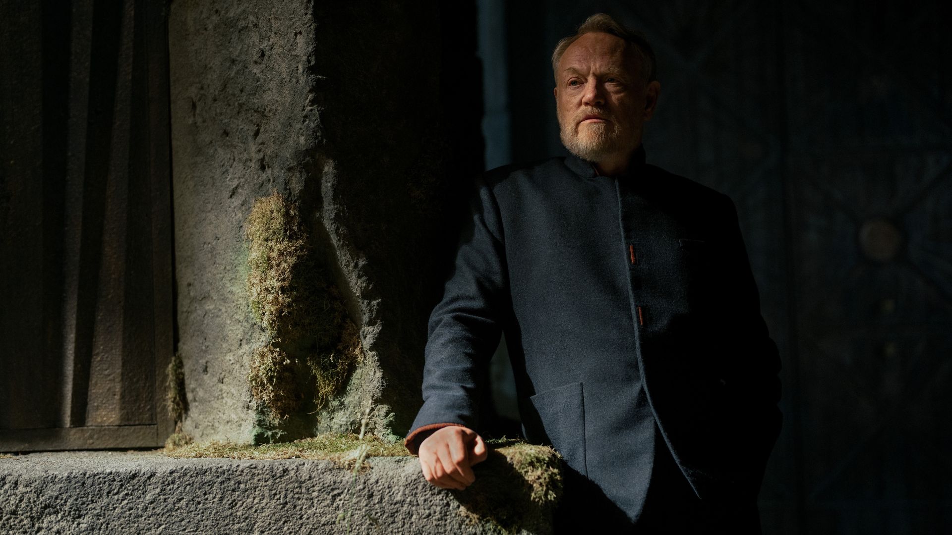 an image of Jared Harris in Foundation. He is wearing a dark suit-like coat and is leaning against an old stone pillar partially covered in moss