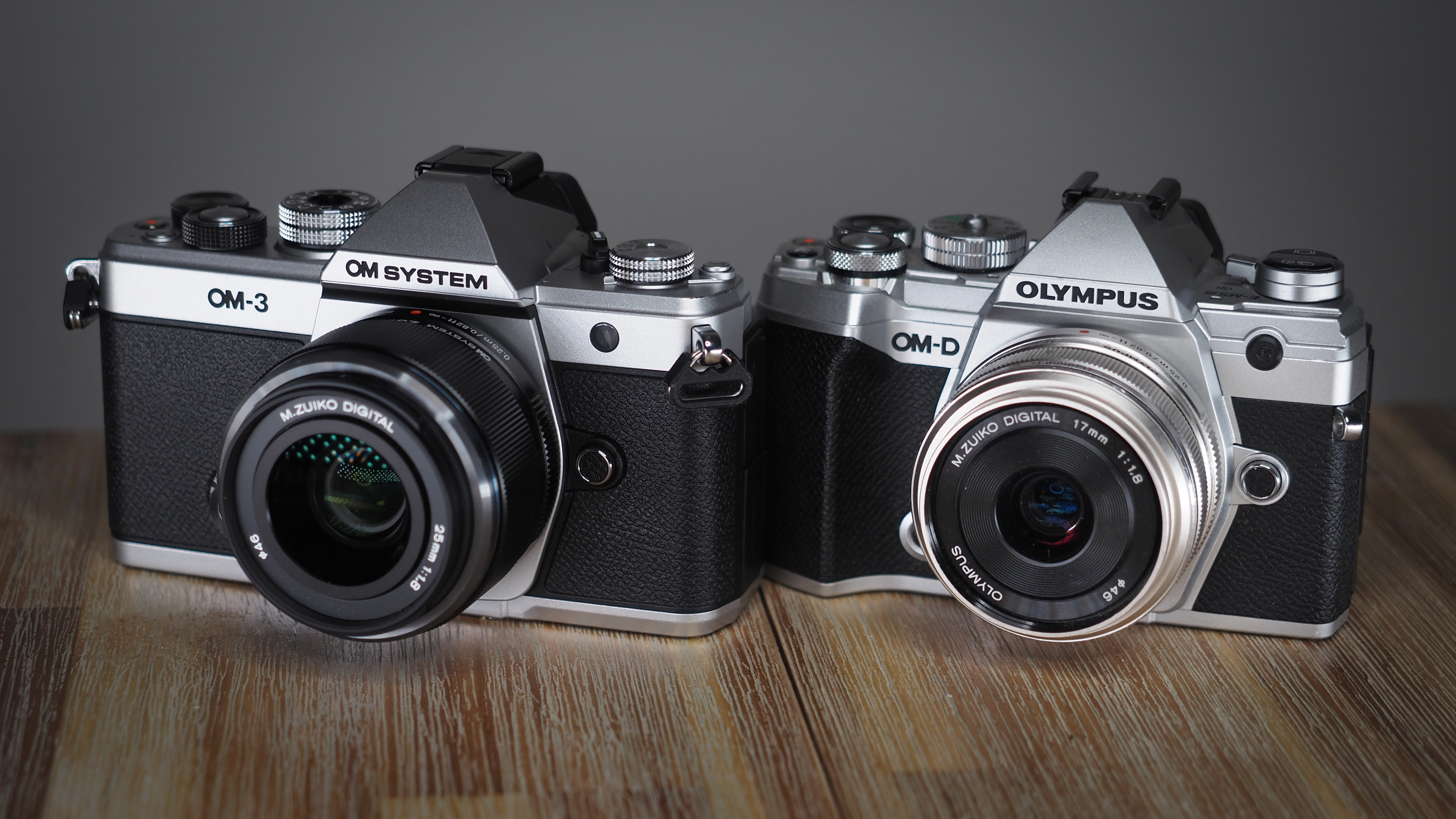 OM System OM-3, next to an Olympus OM-D E-M5 Mark III, on a wooden surface