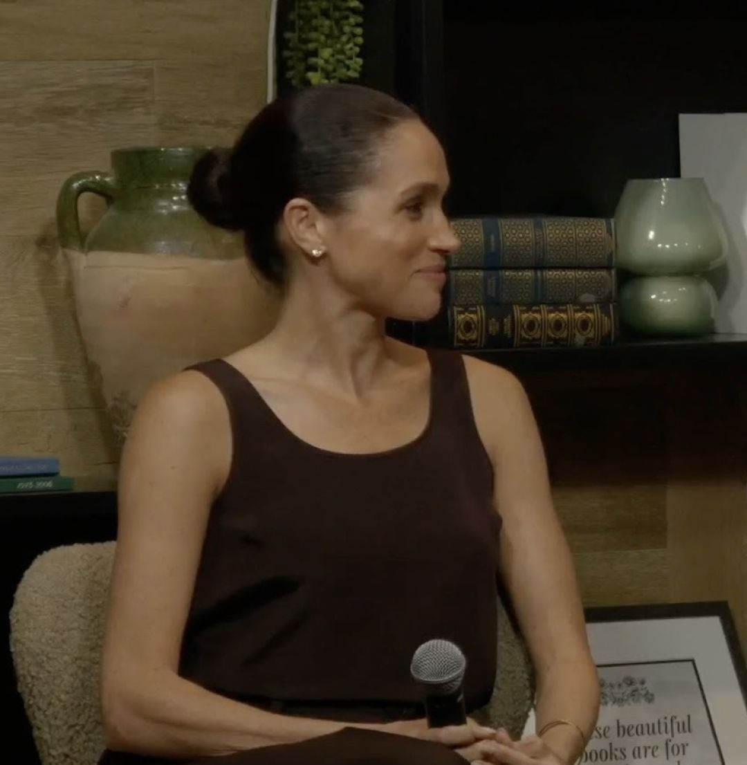 Meghan Markle sitting in a chair wearing a brown tank holding a mic