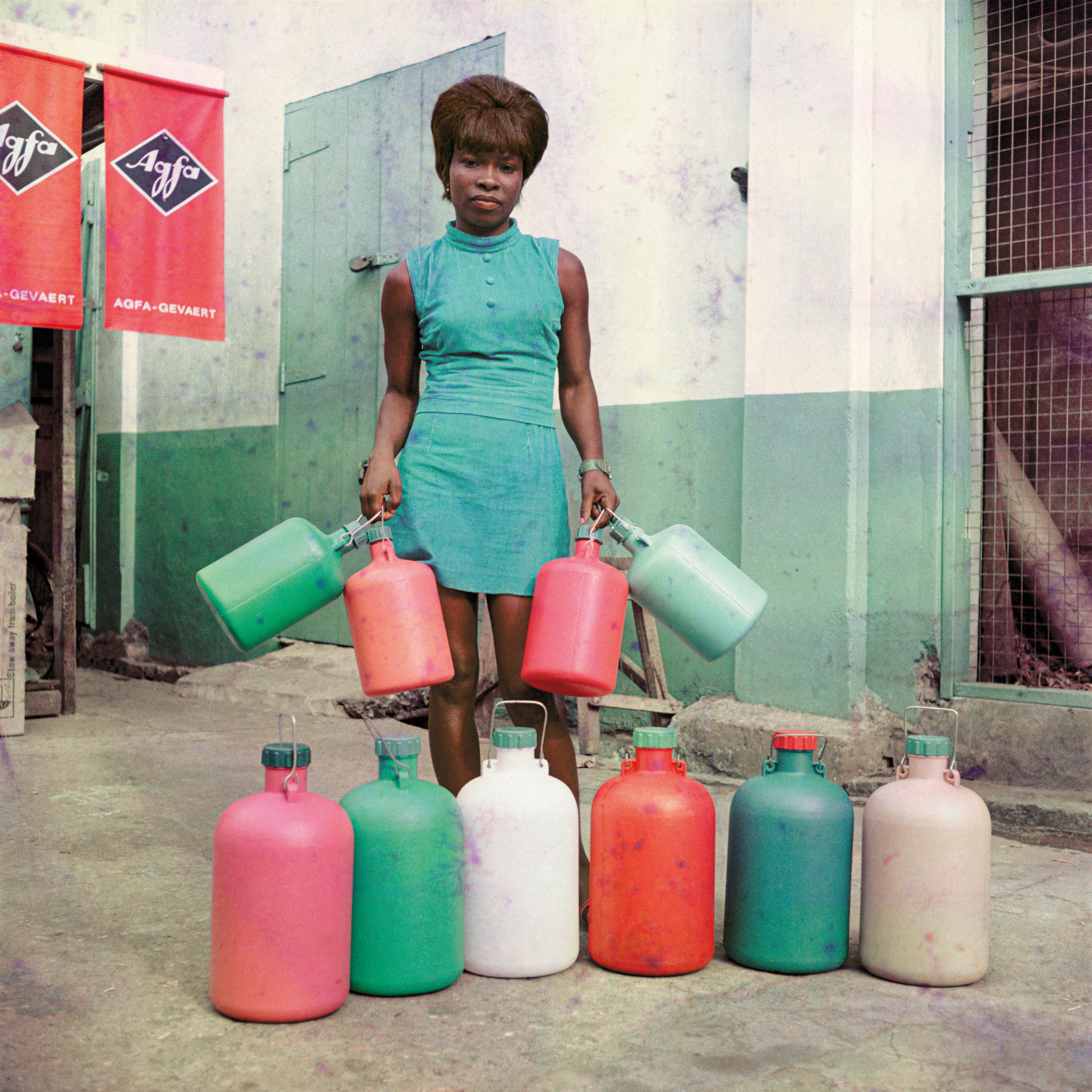 A vibrant color photograph of a woman in a sleeveless blue-green dress holding four large colorful plastic jugs, with six more matching jugs arranged in a line on the ground in front of her.