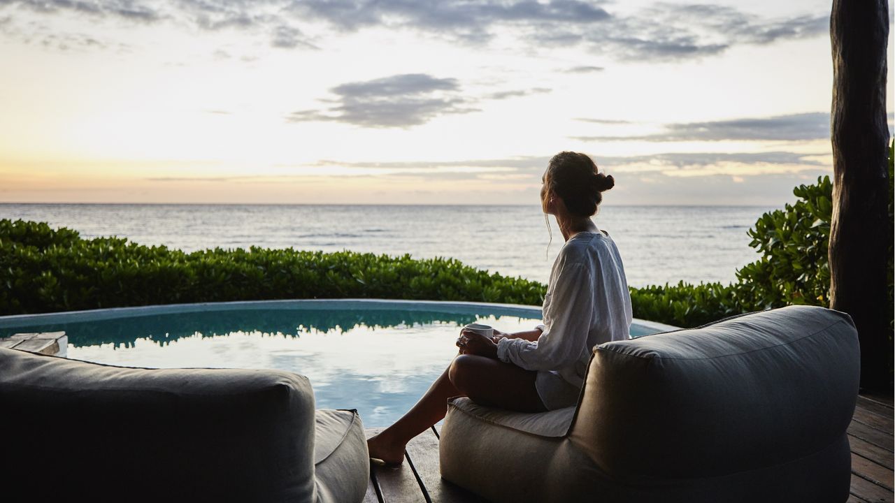 A clearly wealthy woman sits by the pool with a view of the beach.