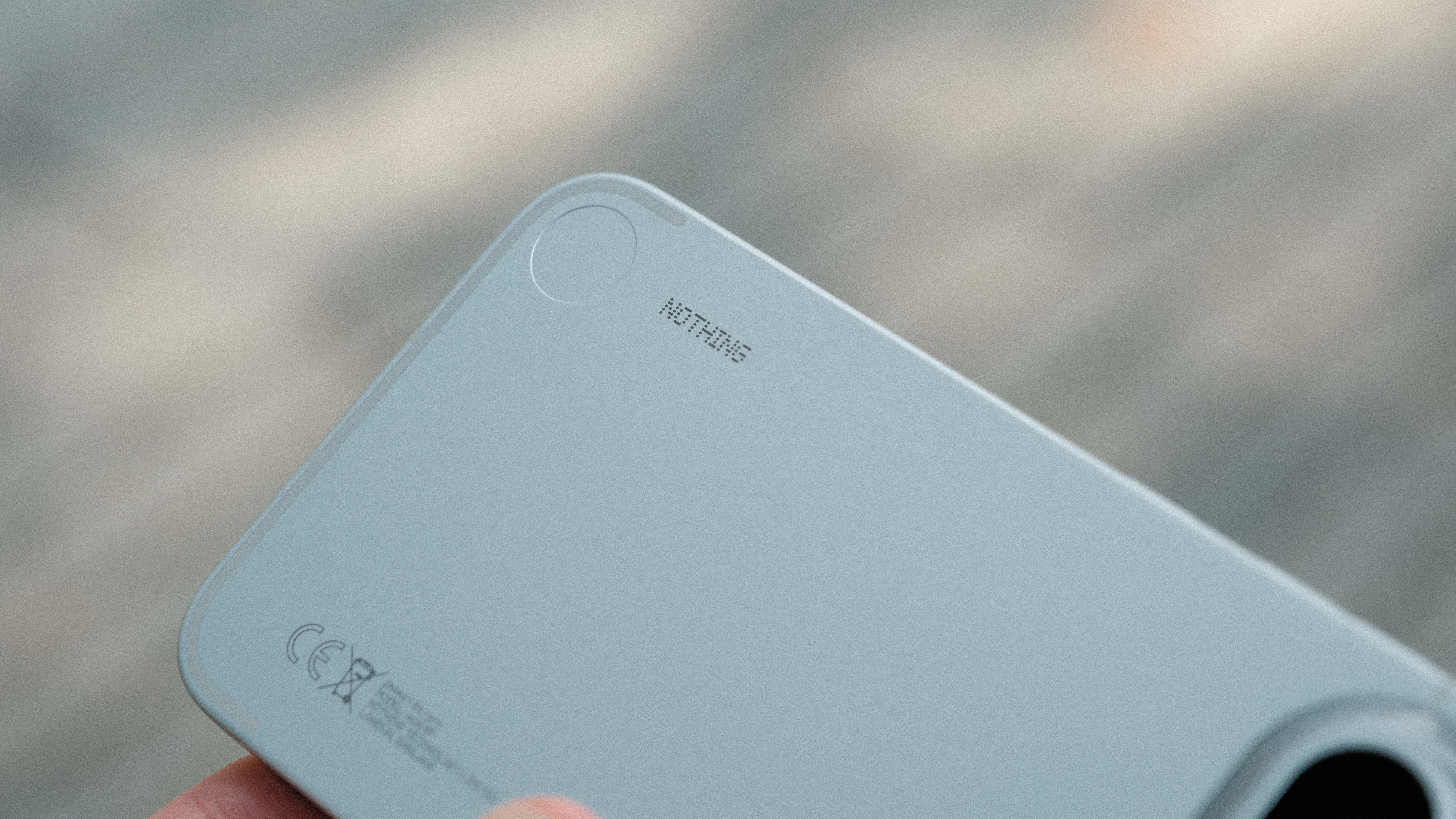 A macro shot of the bottom corner of a matte gray smartphone. The "NOTHING" logo is printed in a signature dot-matrix font near a circular design element, with regulatory symbols and text visible in the lower corner.
