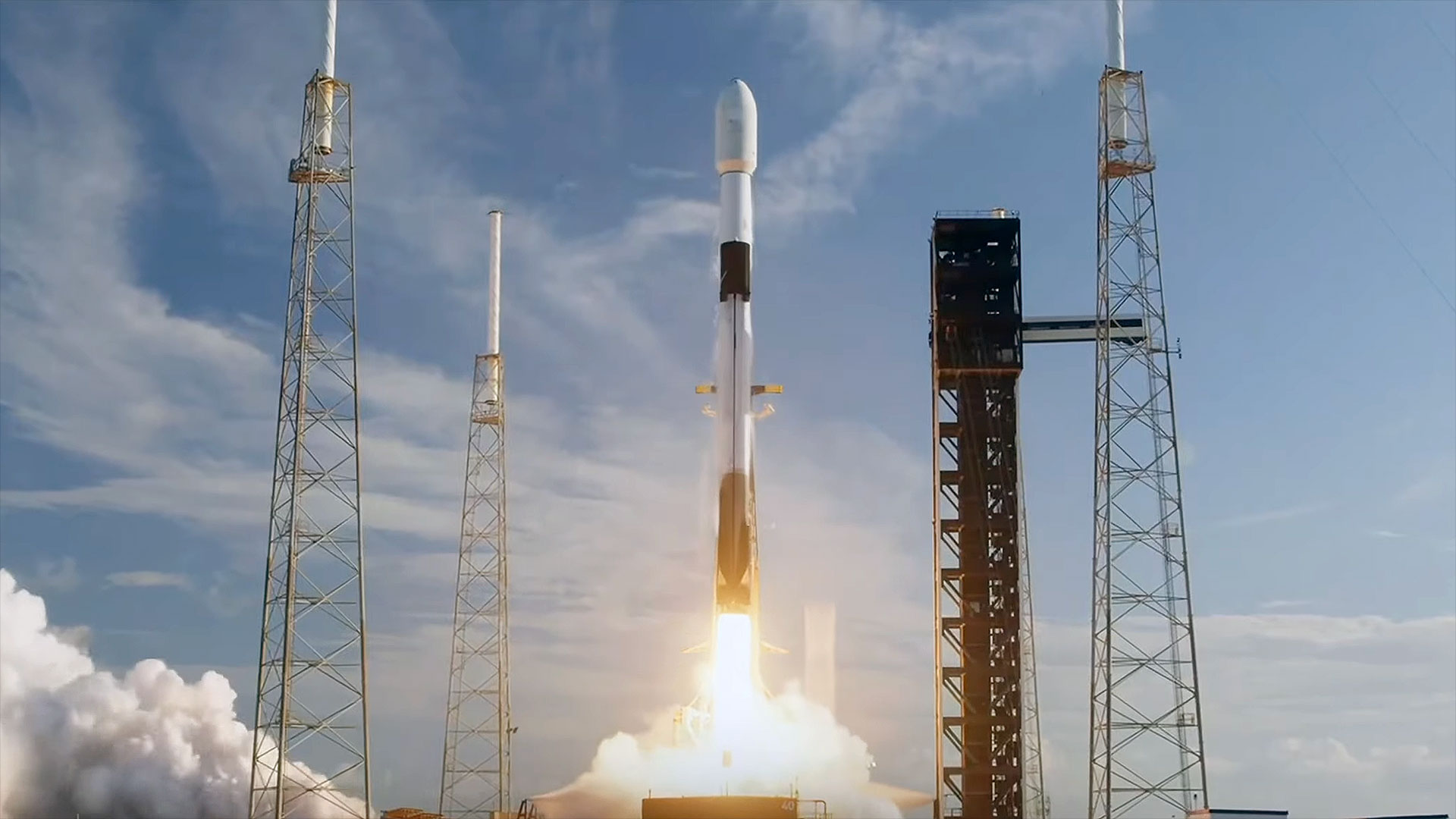 a white and black rocket lifts off on a bright plume with an orange glow, set against a blue sky with wispy white clouds.