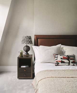 A dark brow padded bed with white bedding and a beige throw. A black bedside table with a black lamp