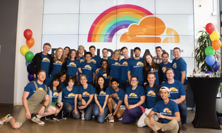 A group of Cloudflare employees wearing t shirts bearing the Cloudflare logo with a rainbow added to the side, againststood and sitting for a photo, against a wall with a large version of the same graphic. To either side of them, a rainbow-assortment of balloons is floating.