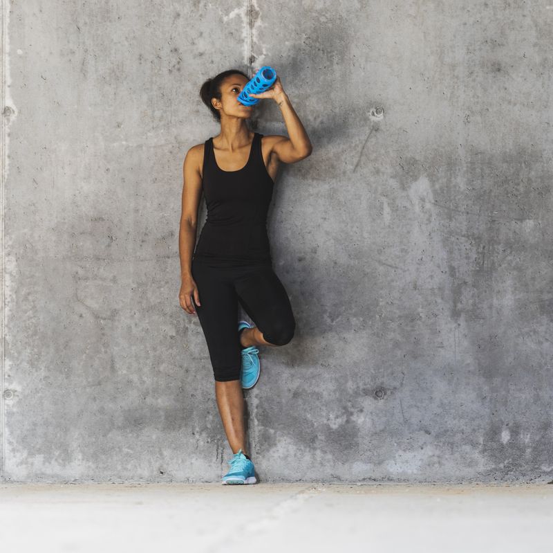 A woman in sports clothing leans against a wall and drinks from a water bottle