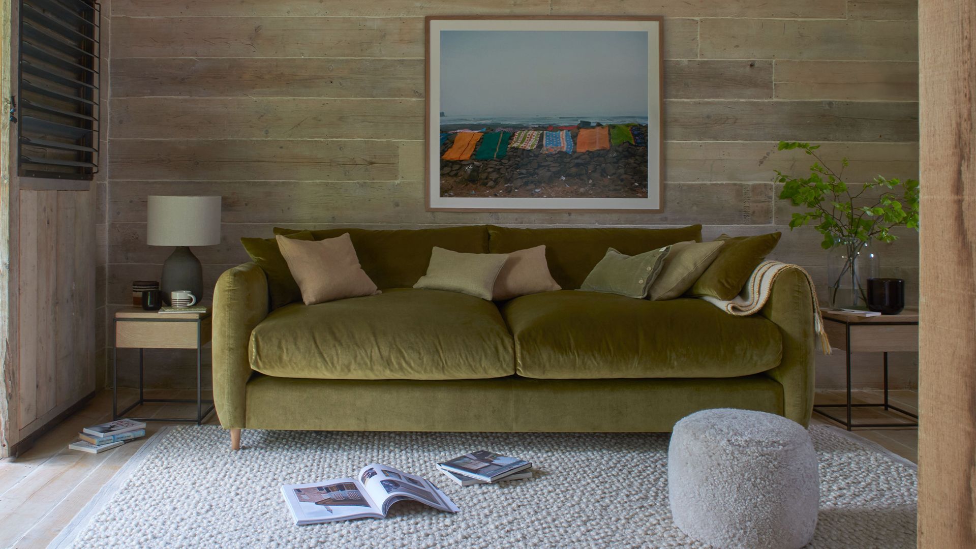 green sofa on a textured fluffy rug with wood textured walls behind and a fabric footstool in front.