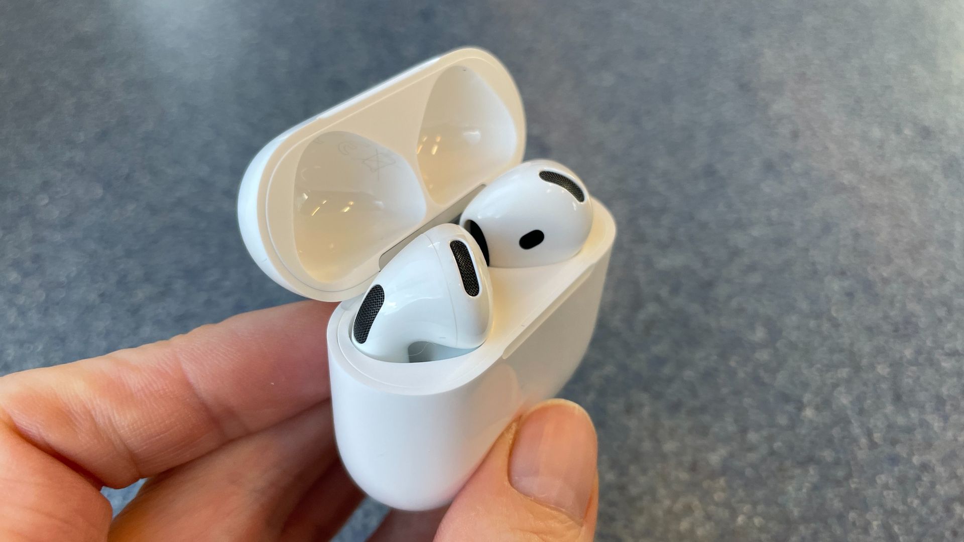 Apple AirPods 5 with Wireless Charging, on a blue table and held in a hand