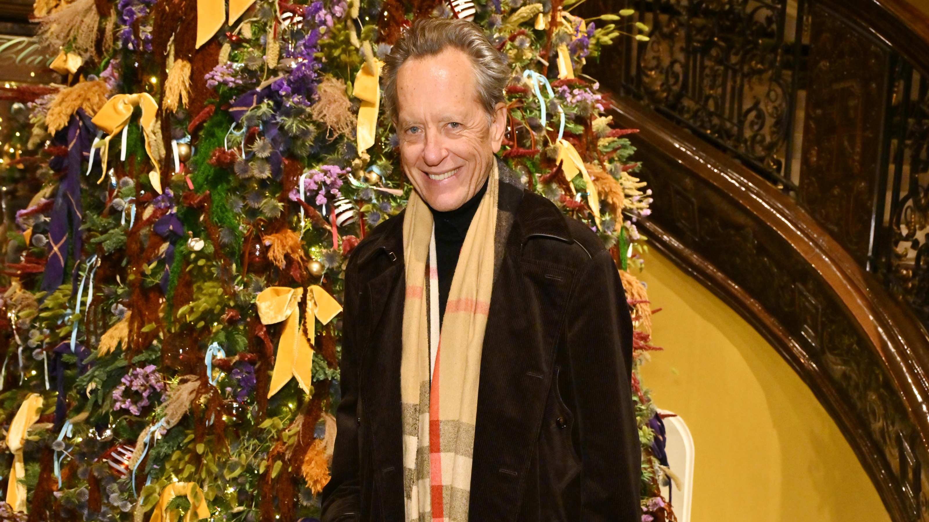 Richard E. Grant wearing a Burberry Scarf stood in front of a large Christmas tree covered in red, purple and mustard bows.