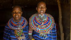 Two Maasai women wearing blue tops with colorful beaded necklaces around their necks 