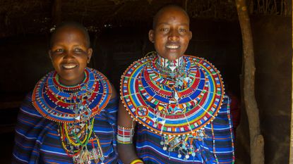 Two Maasai women wearing blue tops with colorful beaded necklaces around their necks 