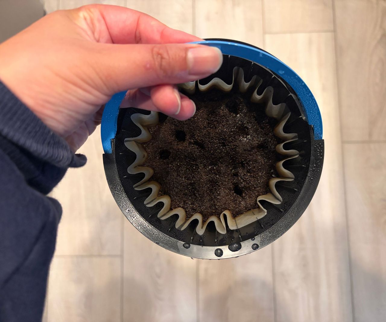 A coffee grinds basket filled with a used filter and grinds, being held aloft.