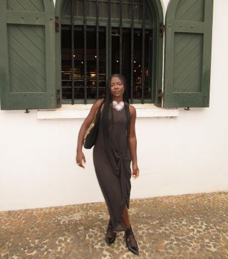 Woman wearing brown pleated front dress, statement necklace, buckled pointy shoes, thin black stockings, standing in front of a white building with green shutters