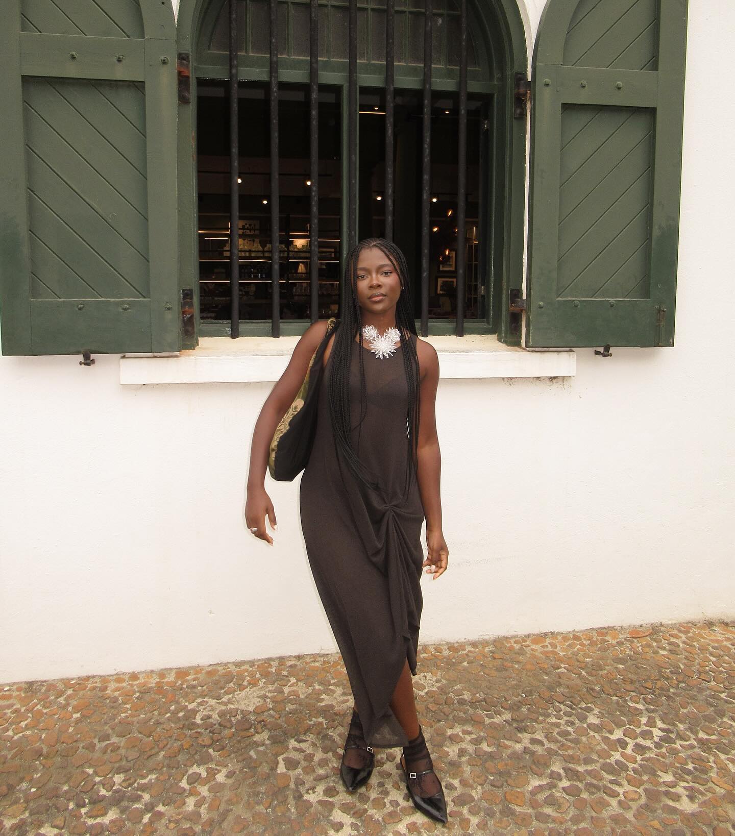 woman wearing brown draped front dress, statement necklace, buckle point-toe shoes, sheer black socks, standing in front of a white building with green shutters