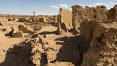 We see sandy-colored rock ruins of old structures in the desert against a blue sky.