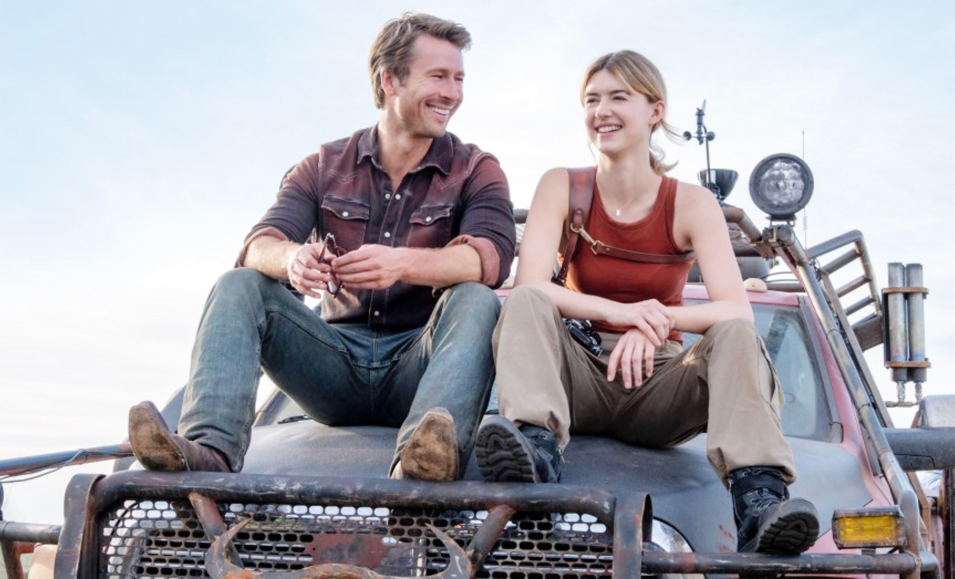 Daisy Edgar-Jones and Glen Powell sit laughing on the hood of a truck decked out in gear