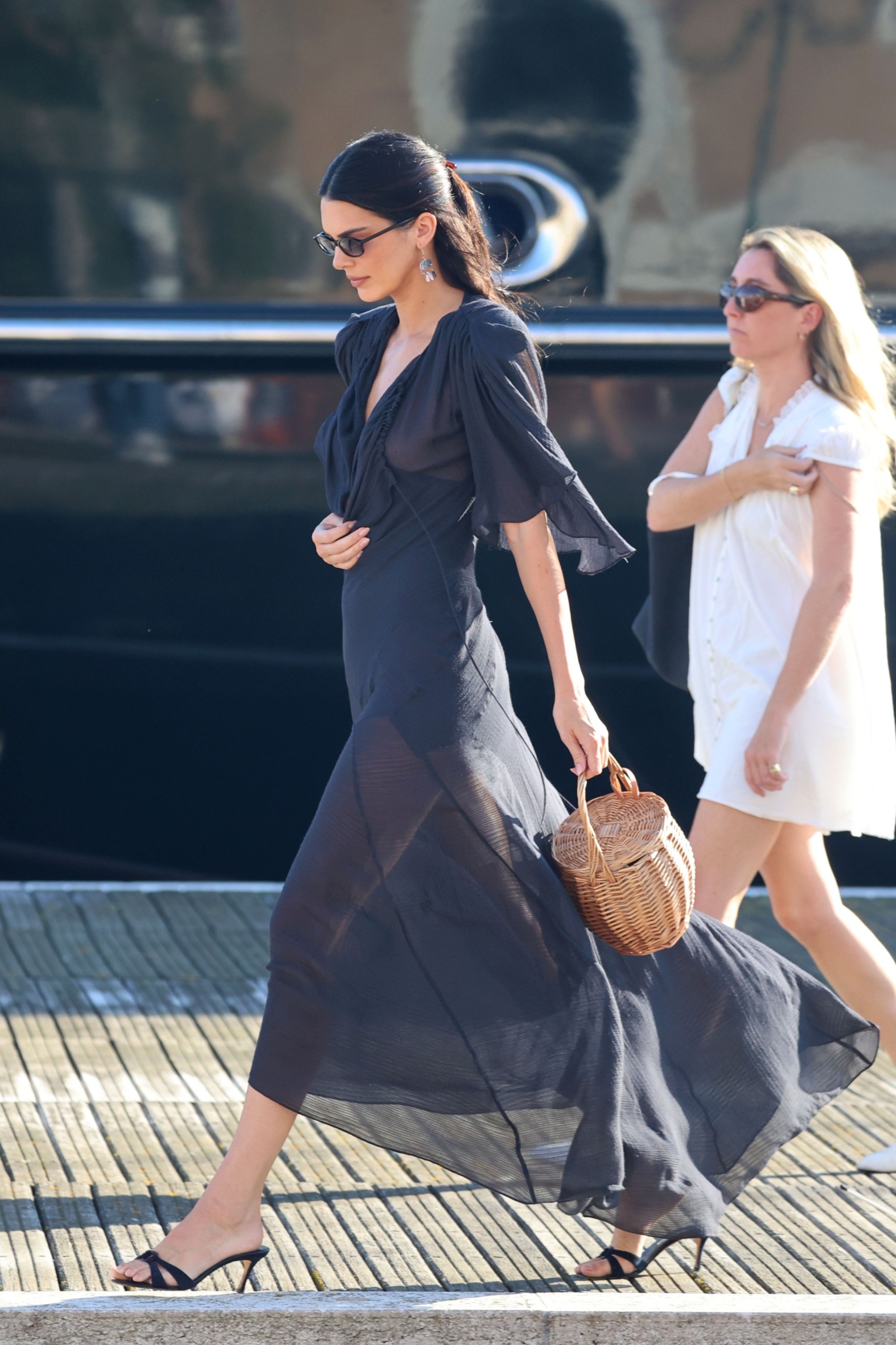 An image of Kendall Jenner wearing a black sheer dress with strappy black mules and a basket bag